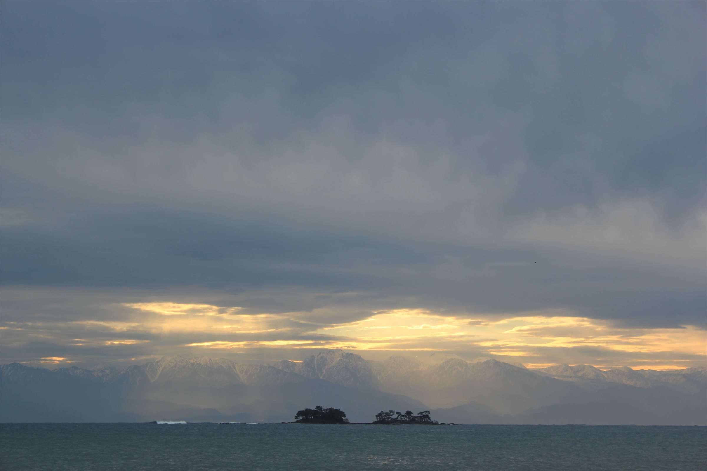 曇り空から差し込む光と立山連峰、小島のシルエット
